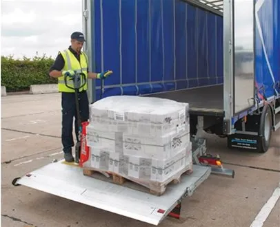 A logistics worker in a high-visibility vest operates a truck’s tail lift, unloading a shrink-wrapped pallet of goods from a blue Palletline trailer in an outdoor loading area.