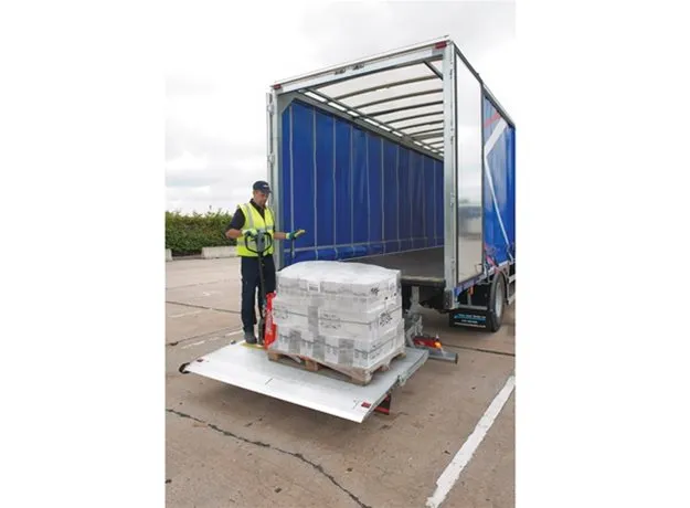 A logistics worker in a high-visibility vest operates a truck’s tail lift, unloading a shrink-wrapped pallet of goods from a blue Palletline trailer in an outdoor loading area.