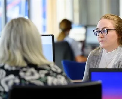 Two women in an office setting, one facing the camera wearing glasses, engaged in conversation with a colleague, with computer screens visible in the foreground.